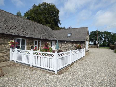 A stone cottage with a gravel driveway and a white picket fence enclosing a garden with flowers and outdoor furniture at Rosemount Coach House in Ballycarney near Enniscorthy County Wexford