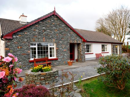 A stone and white cottage with red trim and benches surrounded by flowers and shrubs at The Lodge in Churchtown near Beaufort County Kerry
