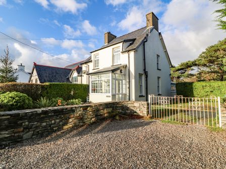 A two-story white house with chimneys and a glass porch behind a stone wall and metal gate at Ty'r Ysgol in Morfa Bychan