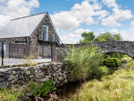 A stone building with a metal fence near a stone bridge over a stream at Glan Nug Bach in Rhydlydan near Betws-y-Coed