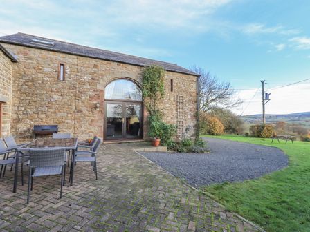 An outdoor patio area with a glass table and chairs beside a stone building at Hadrian's View in Banks near Brampton