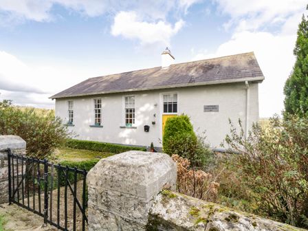 A stone wall and black gate in front of a white house with a yellow door and three windows at The Old School House in Gortermone near Carrigallen County Leitrim