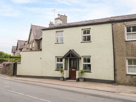 A street view of a two story house with a dark brown door and four windows with two potted plants outside at Mulberry Cottage in Cark in Cartmel