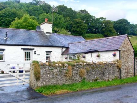 A white stone cottage with a slate roof behind a stone wall and gate in a rural area with trees in the background at Gorrig Bwthyn in Llandysul
