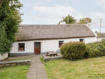 A thatched roof cottage with a wooden door and small windows surrounded by bushes and a picnic table at The Thatched Cottage in Drummin near Westport County Mayo