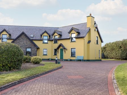 A yellow house with a green door and multiple windows with a stone section on the left and a paved driveway at 4 Golfside in Ballybunion County Kerry