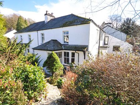 A white two-story house with a slate roof surrounded by bushes and trees at Gavel Cottage in Bowness-on-Windermere