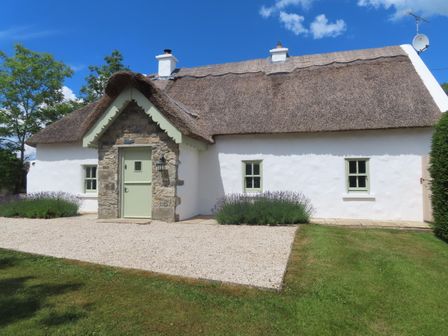 A white cottage with a thatched roof green door stone porch and small windows surrounded by grass and bushes at The Humble Daisy in Woodford County Galway