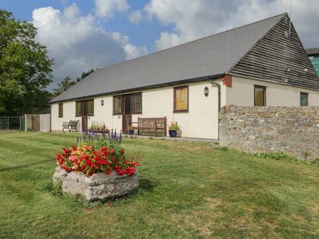 An outdoor area with a long building a stone flower planter and benches at Brindle Cottage in Royal Wootton Bassett