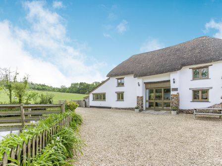 A thatched roof house with gravel driveway and wooden fence surrounded by greenery at Groves Fishleigh in Hatherleigh
