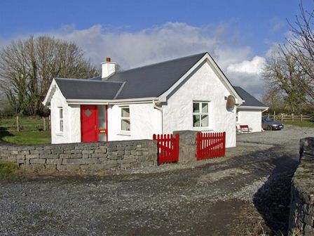 A white cottage with a grey roof red door and gate surrounded by stone walls at Delia's Cottage in Ballinrobe County Mayo