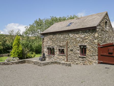 A stone barn with a gravel driveway and garden with trees and chairs at Woodside Barn Pennington near Ulverston