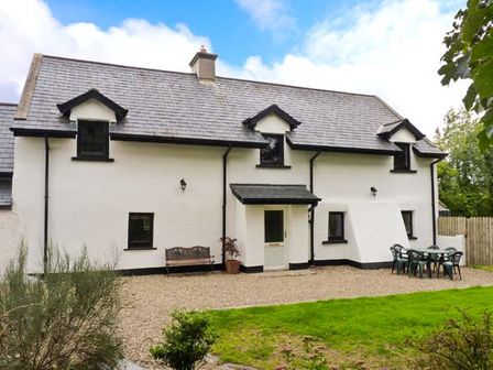 The exterior of a white house with black window frames and a grey roof with a bench a plant and a patio table with chairs in the garden at Home Farm Cottage in Campile County Wexford