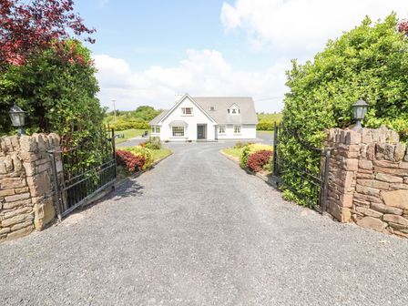 A gravel driveway with open iron gates leading to a white house with a pitched roof surrounded by greenery at Fern View House in Beaufort near Killorglin County Kerry