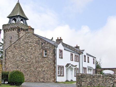 A stone building with a clock tower next to a white house with multiple windows and doors at William Court Cottage Irton Hall near Wastwater