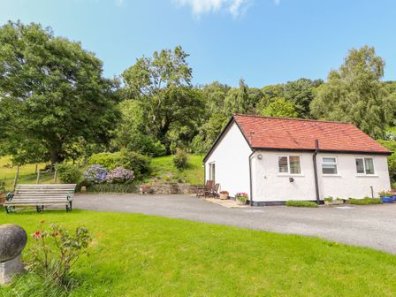A small white house with a red tiled roof next to a paved area and green lawn with trees and a wooden bench at Gwern Tyno in Colwyn Bay
