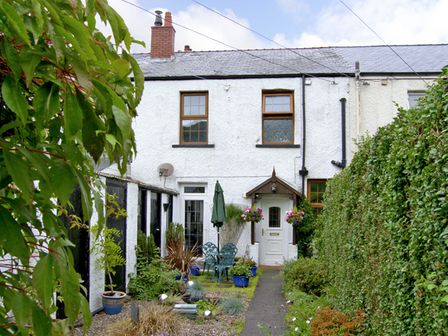 A garden with plants a table chairs and an umbrella in front of a white two story house with wooden windows at Railway Cottage in Fairbourne near Dolgellau