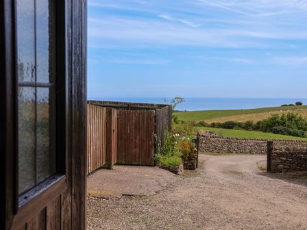 A view of a gravel driveway with a wooden fence and a gate leading to green fields and a stone wall at Dairy Cottage in Staintondale