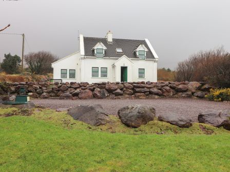 A white house with green trim windows behind a stone wall surrounded by grass at Mount Brandon Cottage in Cloghane County Kerry