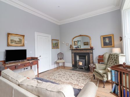 A living room with a beige sofa and armchair a wooden table with a tv a fireplace with a mirror above and paintings on the walls at The Merchant's House in Kilrush County Clare