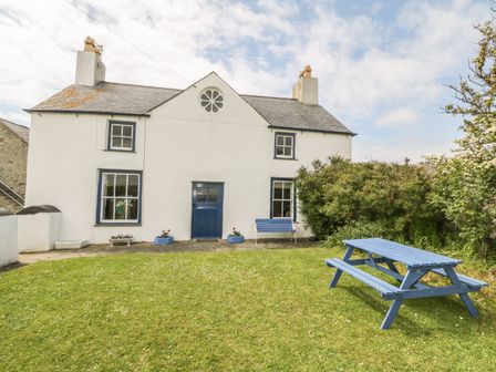 A white two-story house with blue door and window frames a blue bench and a blue picnic table on the lawn at Ty Fferm Bodlasan in Llanfachraeth near Valley