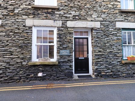 A stone building exterior with a black door and two white window frames on a street at Partridge Holme in Bowness-On-Windermere