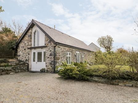 A stone cottage with white doors and windows next to bushes and a gravel driveway at Beudy Hywel in Llanrug near Llanberis