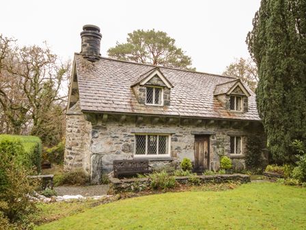 A stone cottage with a tiled roof and wooden door surrounded by trees and grass at Nant Cottage in Llanrwst near Betws-y-Coed