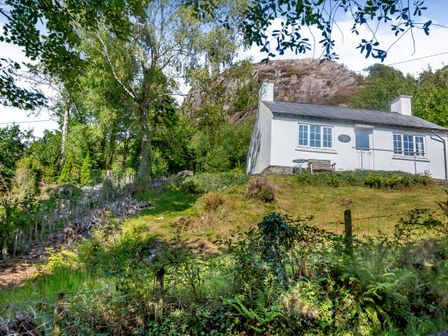 A white cottage on a grassy hill surrounded by trees and a rocky hill behind at Penlan in Beddgelert
