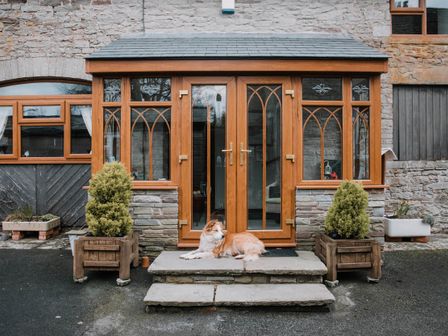 A stone house entrance with wooden framed glass doors and windows a dog lying on the steps and potted plants on each side at Stargazer's Loft in Hardwicke near Hay-on-Wye