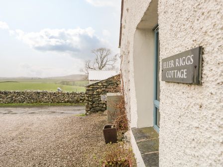 Exterior wall of a cottage with a sign saying Eller Riggs Cottage and a stone wall with fields beyond at Eller Riggs Cottage Eller Riggs Brow near Ulverston