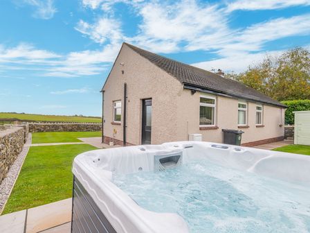 A hot tub in a backyard next to a single-story house with a stone wall and grassy area at Colt Park Cottage in Ulverston