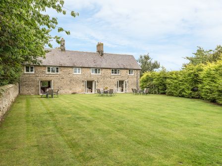 A large grass lawn with outdoor tables and chairs in front of a stone house with multiple windows and chimneys at Stephen's Cottage in Alnmouth