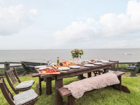 An outdoor dining table set with plates glasses food and a flower vase on grass near the sea at On The Watersedge in Beadnell