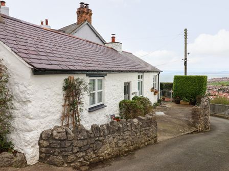 A white stone cottage with a tiled roof and a stone wall in front overlooking a town and sea at Craiglwyd Bach in Penrhynside near Llandudno
