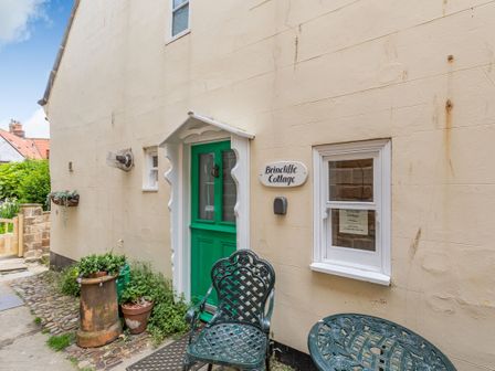 The exterior of a building with a green door a small white window metal garden furniture and a sign reading Brincliffe Cottage in Robin Hoods Bay