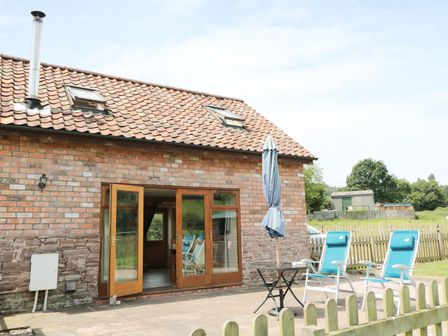 A brick building with a tiled roof and open wooden doors leading to a patio with blue chairs and a closed patio umbrella at Mallard in Newnham-on-Severn near Gloucester