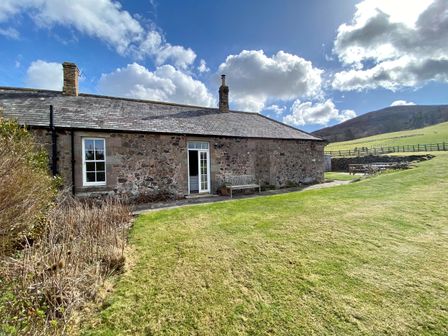 A stone cottage with a bench outside on a lawn with hills and a partly cloudy sky at Akeld Cottage in Akeld near Wooler