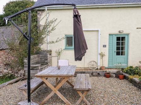 An outdoor patio with a wooden picnic table and benches a closed umbrella and potted plants at Beekeeper's Cottage in Pembroke