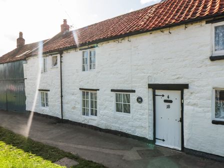 A white stone cottage with a tiled roof and multiple windows along a paved path at Keep Cottage in Pickering