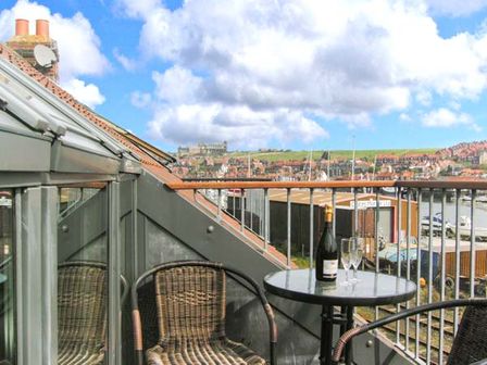 A small balcony with two wicker chairs a round table with a bottle and two glasses overlooking a harbor and buildings at Mariners Rest in Whitby