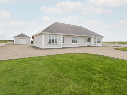 A single-story house with a detached garage and a gravel driveway surrounded by green grass at Shraigh Beach in Belmullet County Mayo