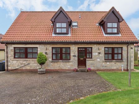 A stone house with a tiled roof and two dormer windows with paved driveway and small tree at Darling Cottage in Bamburgh