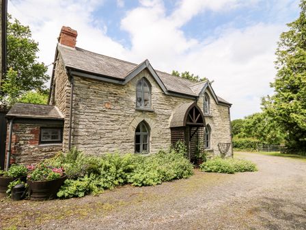 A stone house with arched windows and a wooden covered porch surrounded by plants and gravel driveway at Maesgwyn in Bridell near Cardigan