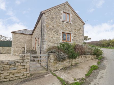 An exterior view of a stone house with wooden windows and a gate by a roadside at The Old Barn in Harman's Cross near Swanage