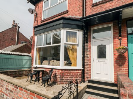 An outdoor patio area with a small black metal table and two chairs next to a white door and a large window with yellow curtains on a brick house
