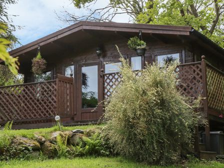 A wooden cabin with a porch and lattice railing surrounded by greenery at Y Caban in Llechwedd The Conwy Valley
