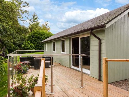 A wooden deck with glass railing and outdoor seating next to a house with green siding at Solway Cottage in Bowness-on-Solway near Wigton