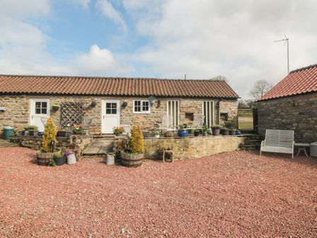 An outdoor area with a stone building with tiled roof multiple white doors and windows a raised stone patio with potted plants chairs and a bench on gravel at Norah's Nook in Farndale near Kirkbymoorside