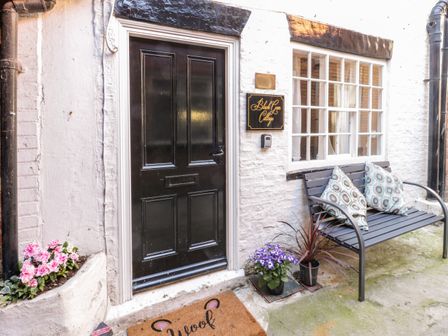 The entrance of Black Gem Cottage in Whitby with a black door a window a bench with two patterned cushions and potted flowers